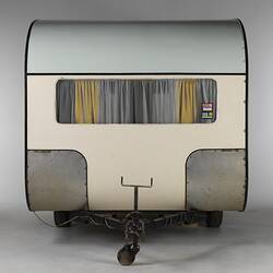 Cream caravan, curved edges, metal trim, front view. One window, yellow white curtain. Tow bar.