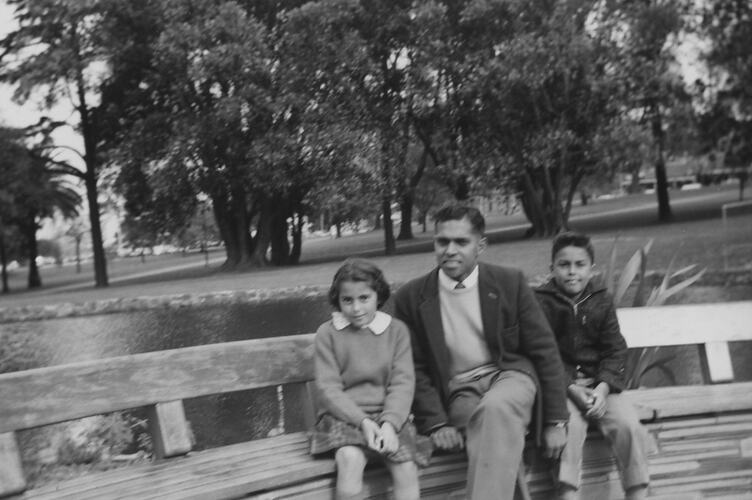 Man and two children sitting on park bench in a park.