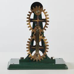 Model of two brass coloured metal elliptical gear wheels on a frame with a handle, mounted on green base.