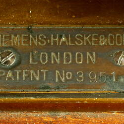 Brass apparatus with batteries on wooden base, detail of label.