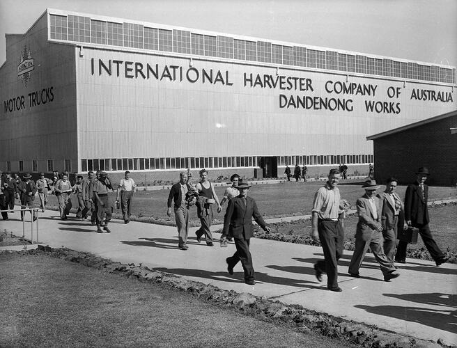 Men leaving a factory via a concrete path. They wear jackets, hats or caps, and some carry Gladstone bags.