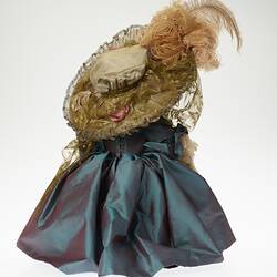 Back of teddy bear wearing lavish cream hat with feather and blue dress jacket.