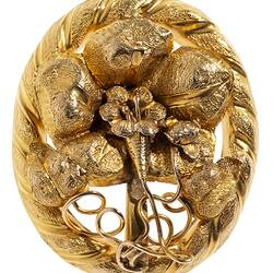 Oval decorative gold brooch containing a flower inside a larger flower, all within an oval border entwined with gold ribbon.