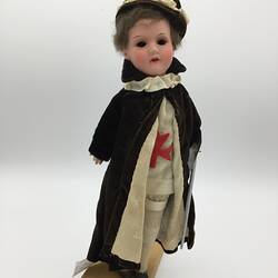 A doll dressed in a cream tunic with a red cross, cream pants and a dark cloak