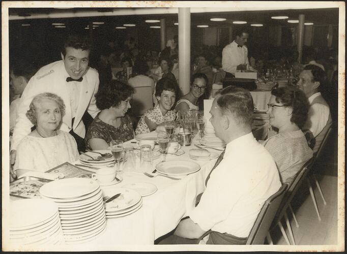 Family seated at a dining table. Some members and the waiter smile at the camera.