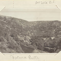 Sloping, scrub covered landscape with two tents and truck on the right bottom side.