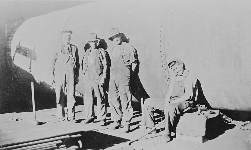 Four men in front of boiler. Man at right is seated.