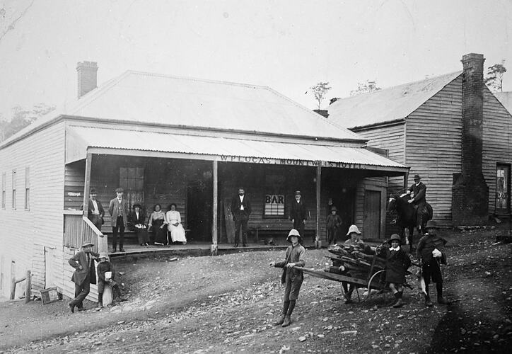 People in front of a hotel with a veranda. A boy is pulling a wood-laden cart at the right.