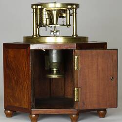 Brass clock under glass dome, supported by wooden octagonal box with door. Cabinet door open showing brass tap