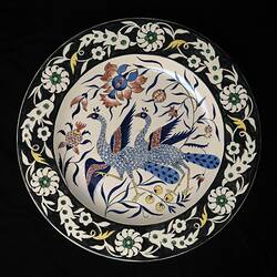 Plate painted with two blue peacocks and flowers. Black rim has white floral motif. Black background.