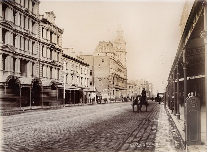 Several horse drawn vehicles on a cobblestone street, buildings either side.