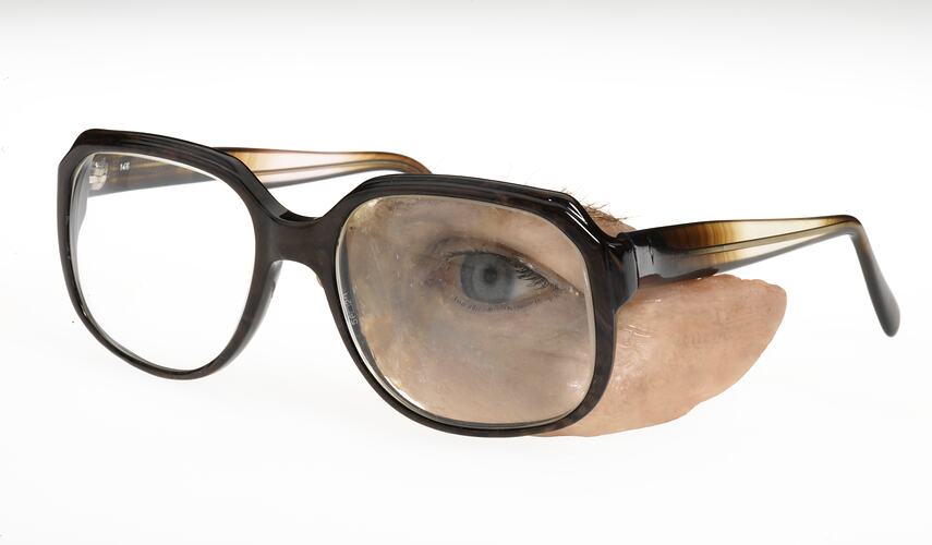 Plastic prosthetic glass blue/grey left eye with upper left cheek, eyebrow and eyelashes attached to brown plastic spectacles. Left front angle view.