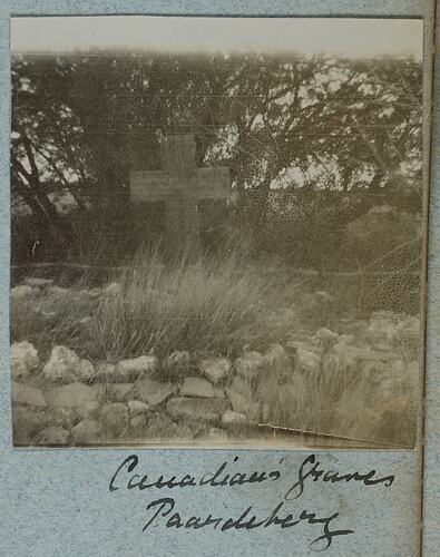 Grave with cross head stone, with stones and bushes in front and trees behind.