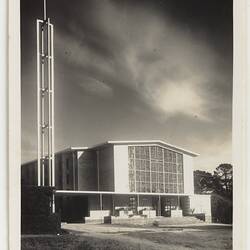 Exterior of white church with large central rectangular glass. White cross on metal tower at left.