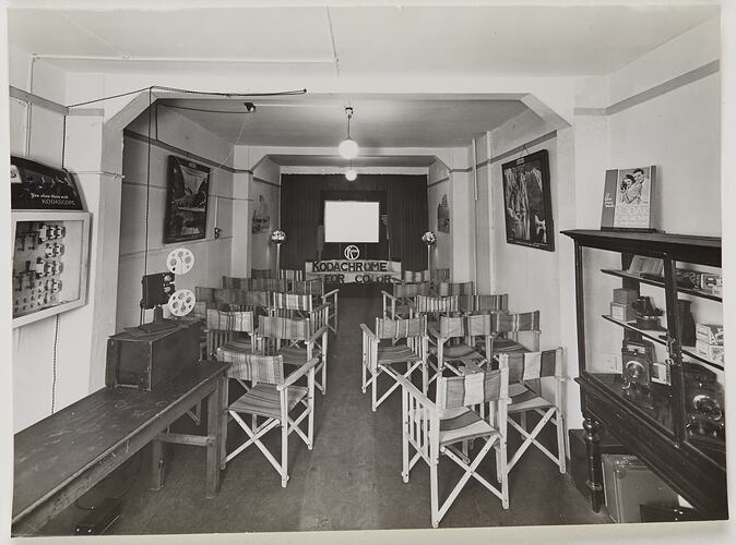 Interior of room set up with folding wooden chairs that face a screen at the front. Display cabinet at right h