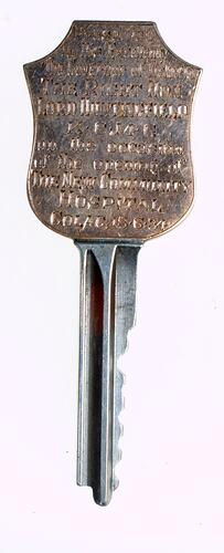 Metal key with silver shaft and gold shield-shaped bow that is engraved.