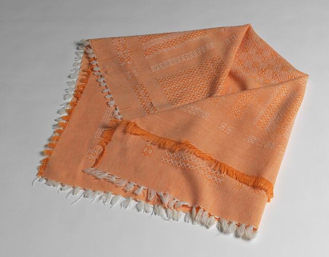 Folded orange and white woven baby blanket with geometric patterns and orange and white fringe around edges.