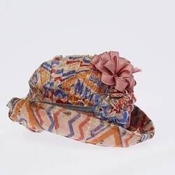 Doll sized hat made from colourful lolly wrappers. Oval shaped with deep upturned brim. Flower on top.