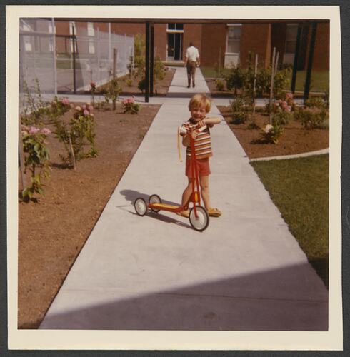 Boy posing on a footpath outside a building with a scooter. He wears red.
