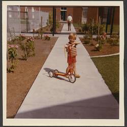 Boy posing on a footpath outside a building with a scooter. He wears red.