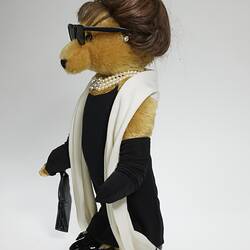 Profile, brown bear wears black hat, sunglasses, dress, full-length gloves, bag, shoes, white pearls and scarf