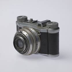 Black and silver camera. Central lens at front and two dials on top. Three quarter view.