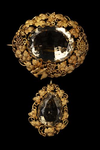 Two part brooch. Top, ornate gold frame with transparent brown cut stone. Below hangs smaller teardrop version