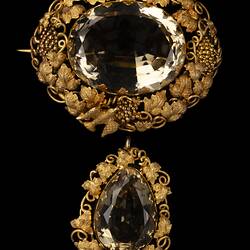 Two part brooch. Top, ornate gold frame with transparent brown cut stone. Below hangs smaller teardrop version