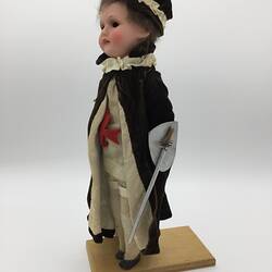 A doll dressed in a cream tunic with a red cross, cream pants and a dark cloak