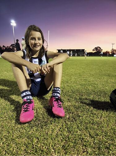 Girl sitting on football ground wearing pink footy boots and black and white striped football jumper and dark shorts. Dusklit horizon.