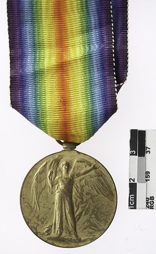 Round gold coloured medal with winged woman and rainbow ribbon attached.