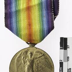 Round gold coloured medal with winged woman and rainbow ribbon attached.