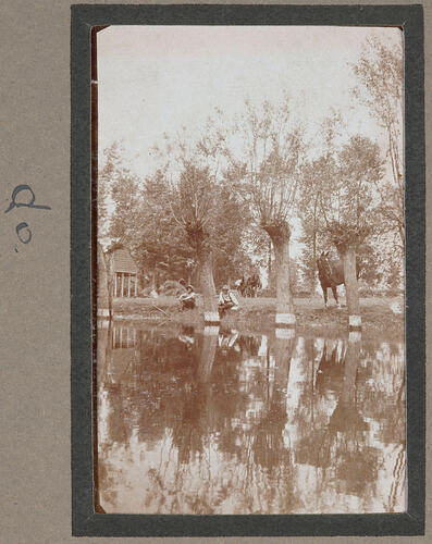 Small pond surrounded by leafless trees, two men sitting on edge of water and two horses grazing behind.