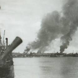 Part of sailing ship on left, land, buildings and billowing smoke in the background.