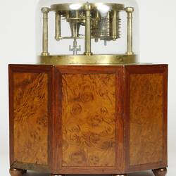 Brass clock under glass dome, supported by wooden octagonal box with door. Back view.