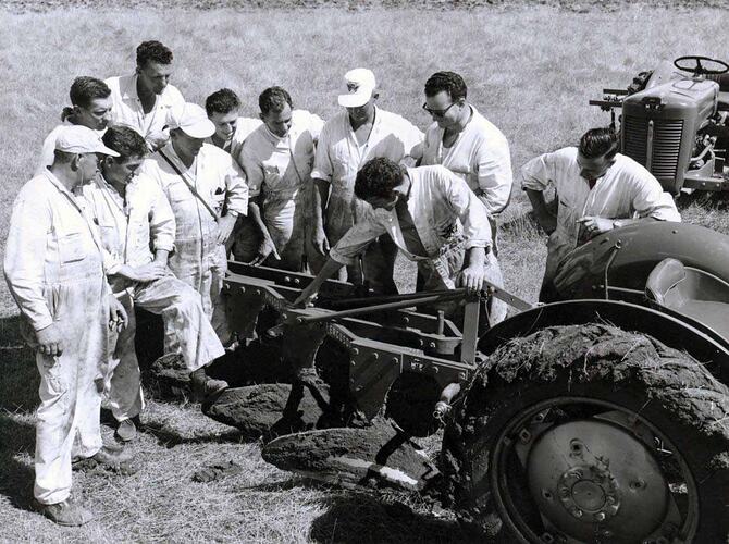 Group of men stand beside tractors in field.