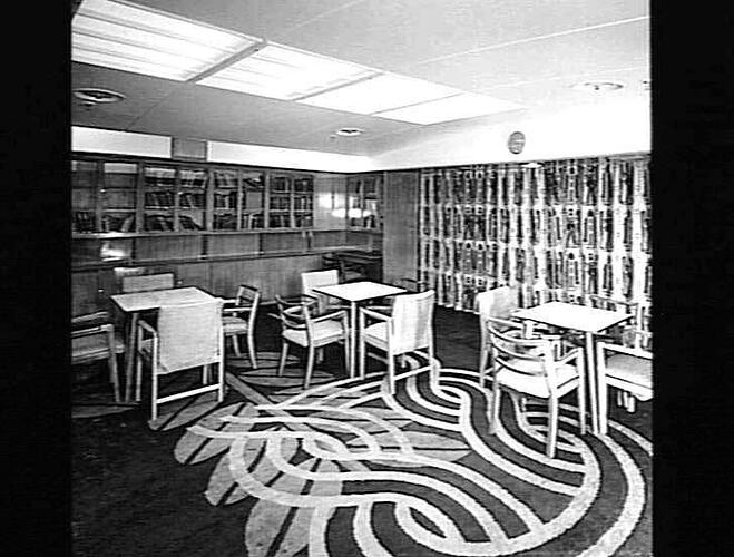 Ship interior. Library shelf on back wall. Some chairs and tables. Patterned carpet.