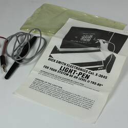 Black pen with grey cable attached, white page with black printing. Plastic sleeve.