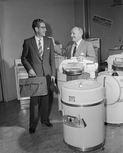 Two men standing with a washing machine. One man holds a hat and satchel bag. Second man has hand on washer.