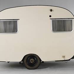 Cream caravan, curved edges, metal trim. Two side windows. Has two wheels and a tow bar.
