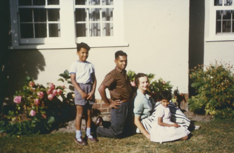 Man kneeling, woman seated and two children outside a house.