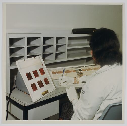 Woman making notes on prints, seated at desk.