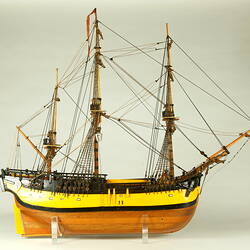 Model ship with wooden hull painted yellow and three masts, facing right.