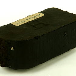 Rectangular black briquette with broken end and paper label.