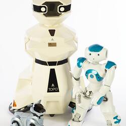 Cream plastic robot with angular human-like shape, ie. head, shoulders, waist and legs. Siver robot dog at left, smaller blue white root at right.