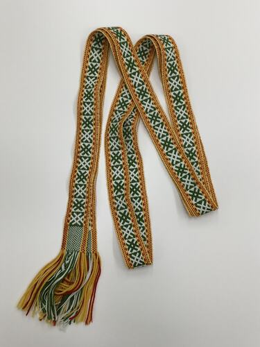 Long woven belt, cream with green criss-cross design. Gold, red border along edges. Fringed ends.