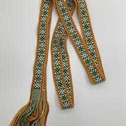 Long woven belt, cream with green criss-cross design. Gold, red border along edges. Fringed ends.