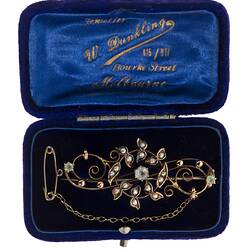 Central gold circle of seed pearls overlaying gold scroll with three paste stones. Fine chain and pin at right. In blue box.