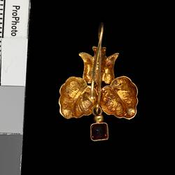 Back of gold pendant earring with downward facing bird, two leaves and single red hanging charm.