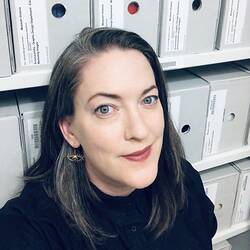 A pale-skinned woman with dark straight hair smiling in front of racks of archive boxes.